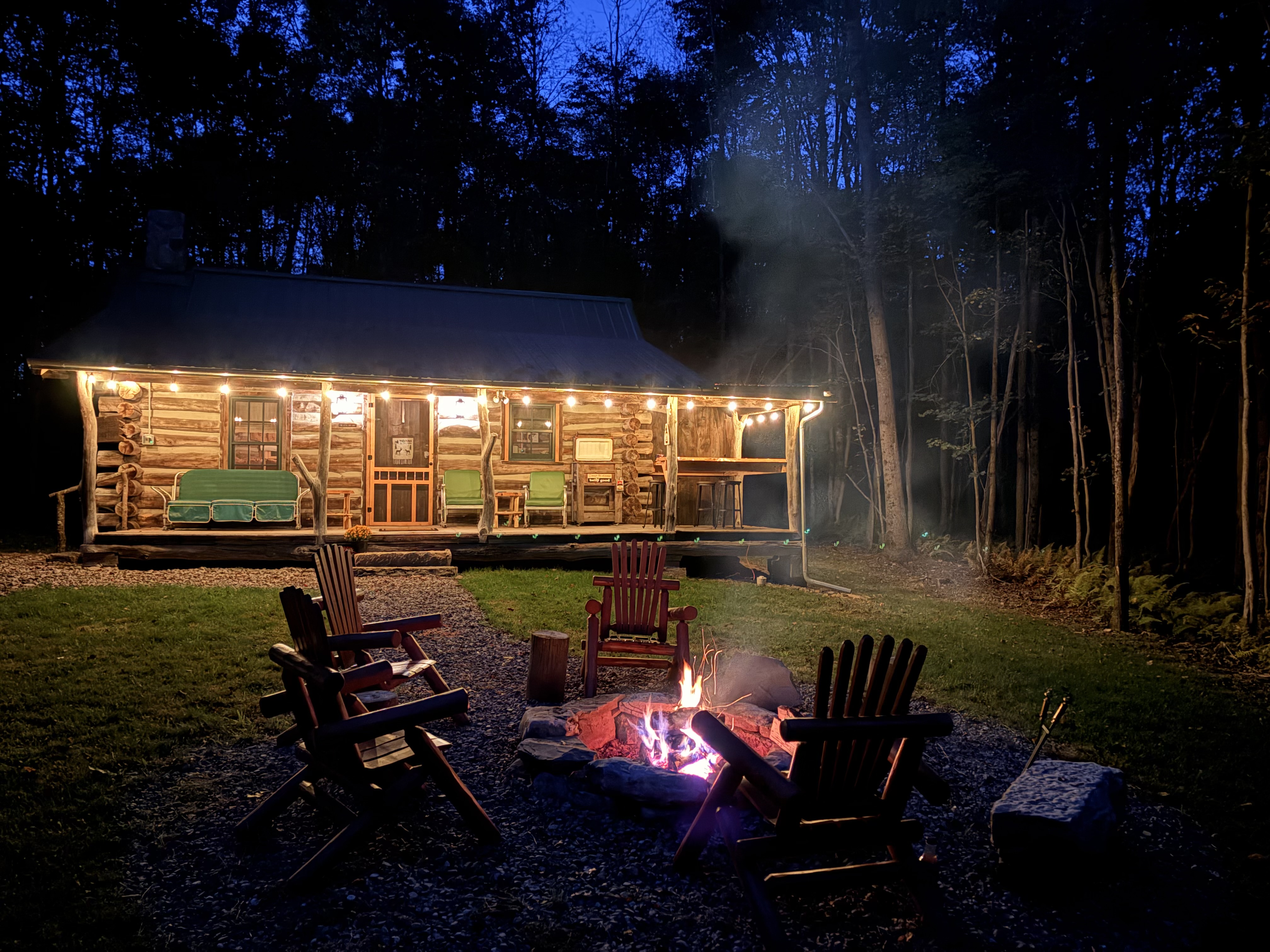 Hogback Log Cabin at dusk, with glowing porch lights and a fire in the outdoor fire pit